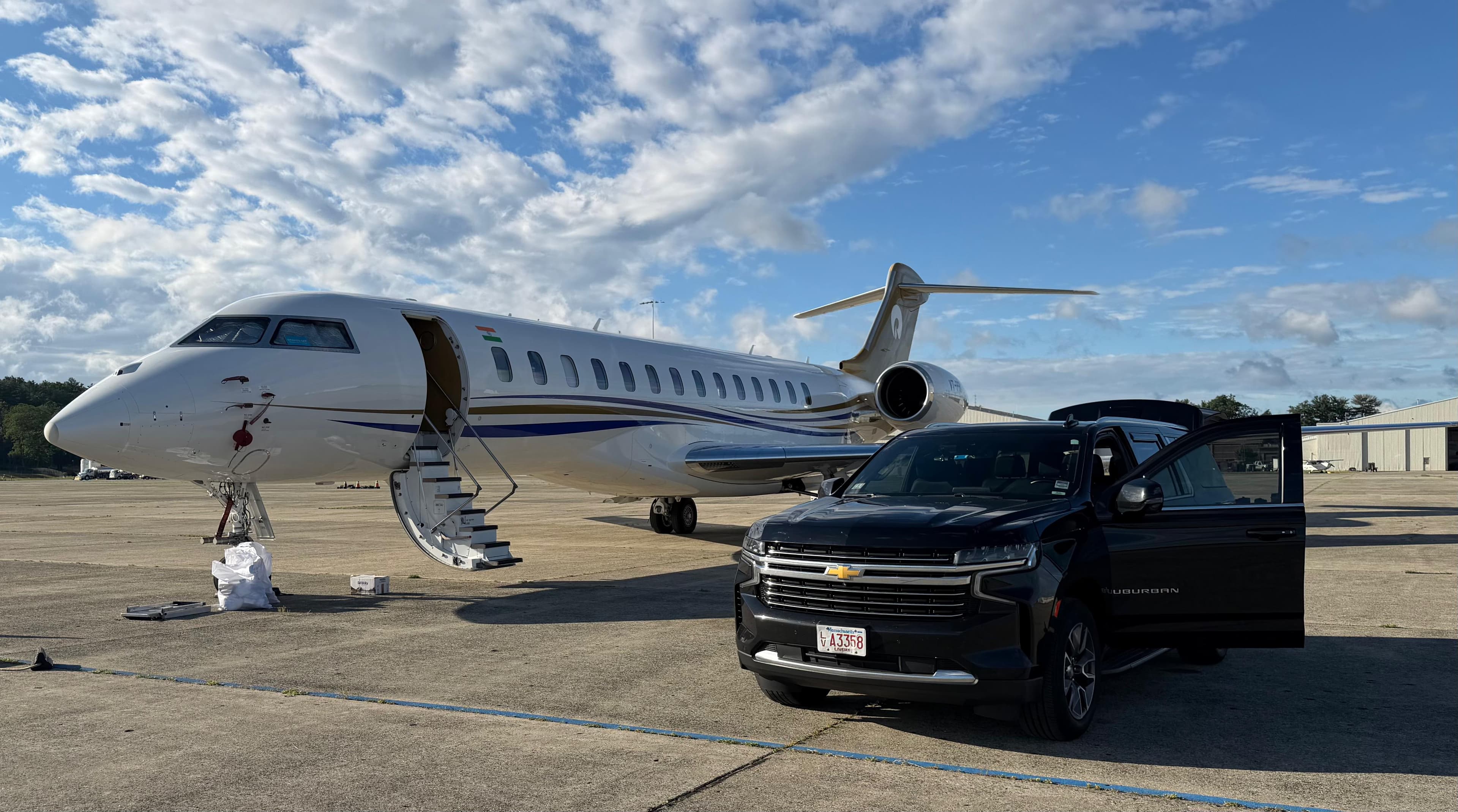 White private jet with stairs down beside a black SUV on a sunny airport tarmac.
